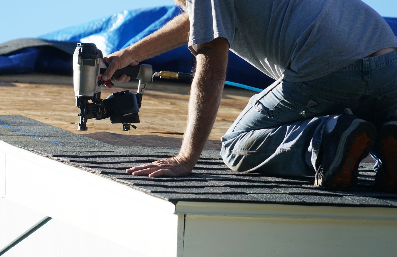 Worker using nail gun on roof shingles for home construction.